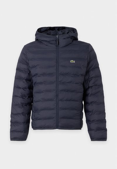 Doudoune matelassée bleu foncé avec capuche et fermeture éclair à l'avant, avec un petit logo crocodile vert sur le côté gauche de la poitrine.