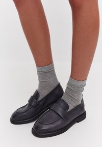 Zwarte leren loafers met een platte zool en een grote decoratieve riem, gecombineerd met grijze geribbelde sokken. Focus op minimalistisch ontwerp en comfort.