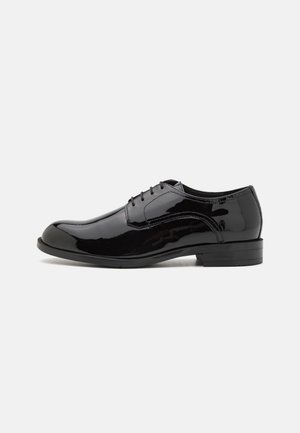 Chaussure habillée pour homme en cuir verni noir avec fermeture à lacets et talon bas sur fond blanc.