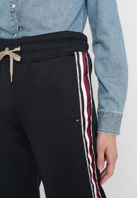 Černé tepláky s ribbovaným pasem, zlatou šnůrkou a vertikálními pruhy v červené, bílé a béžové barvě po stranách. Texturovaná tkanina.