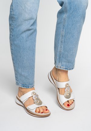 Füße, die weiße Sandalen mit dekorativen Perlen und silbernen Kreisen tragen, kombiniert mit hellblauen Jeans vor schlichtem Hintergrund.