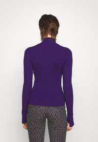 Lila ribbad stickad tröja med hög krage och figursydd design, bärs med grå leopardmönstrade byxor. Texturerat tyg och långa ärmar synliga.