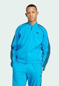 Tuta sportiva blu realizzata in materiale liscio, con zip frontale, strisce nere di accentuazione sulle maniche e logo vicino al petto e alla gamba dei pantaloni.
