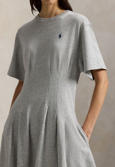 Γυναίκα που φοράει γκρι φόρεμα με κοντά μανίκια, τσέπες στα πλάγια και μικρό κεντητό λογότυπο σε ναυτικό μπλε στο στήθος.