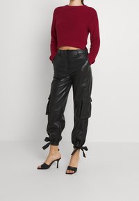 Lyhyt, punainen neulottu villapaita, jossa on pitkät hihat, paritettu mustiin nahkakarhurihmavaatteisiin, joissa on taskut ja säädettävät nauhat hihansuissa.