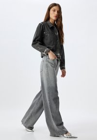 Chaqueta de mezclilla negra con bolsillos delanteros y botones plateados, combinada con jeans gris de pierna ancha de cintura alta. El modelo lleva zapatos plateados con punta.