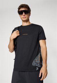Man draagt een zwart "icebreaker" t-shirt met een contourpatroon aan de zijkant, grote zwarte zonnebril en een zwarte rugzak over één schouder.