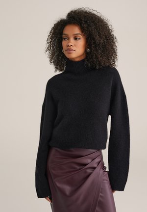 Sieviete ar cirtainiem matiem, ģērbta melnā turtleneck džemperī un tumši purpursarkanā ādas svārku fons ir vienkāršs pelēkbrūns.
