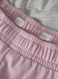 Ceinture élastique rose réglable avec des boutonnières et des boutons blancs sur un fond de tissu gris clair.