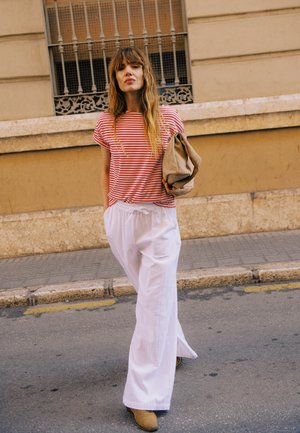 Femme marchant dans la rue portant une chemise rayée rouge et blanche, un pantalon blanc à jambes larges, des bottes beige et tenant une pochette beige.