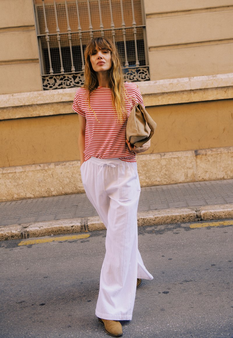 Femme marchant dans la rue portant une chemise rayée rouge et blanche, un pantalon blanc à jambes larges, des bottes beige et tenant une pochette beige.