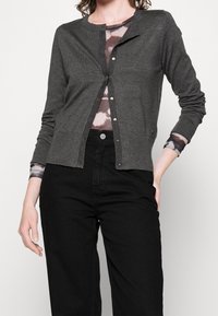 Szary sweter z dzianiny z okrągłym dekoltem, guzikami z przodu i ściągaczami przy mankietach. Nosi się go na koszuli z wzorem tie-dye i łączy z czarnymi spodniami.