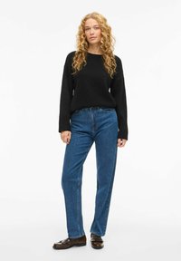 Ung kvinna med lockigt blont hår iförd en svart tröja, blå raka jeans och mörkbruna loafers, stående mot en vit bakgrund.