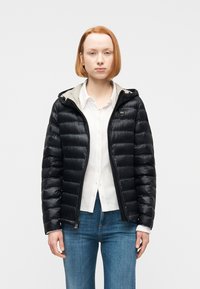 Νεαρή γυναίκα με κοντά κόκκινα μαλλιά, ντυμένη με μαύρο μπουφάν τύπου puffer, λευκό πουκάμισο με κουμπιά και μπλε τζιν, που στέκεται μπροστά σε απλό φόντο.