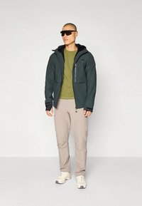 Donkergroene capuchonjas met ritssluitingen, gecombineerd met een lichtgroen shirt en beige broek. Model draagt een zonnebril en witte sneakers.