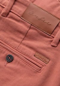 Roter Baumwollstoff mit strukturierter Oberfläche, ausgestattet mit einem Leder-Patch und einem schwarzen Knopf. Enthält genähte Details und ein kleines Etikett.
