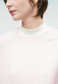 Maglione rosa chiaro con colletto e polsini a costine, realizzato in materiale morbido. Indossato sopra un dolcevita bianco, che mette in evidenza una texture liscia.