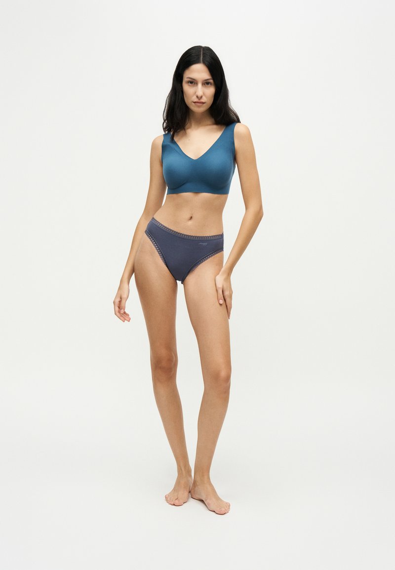 Femme aux longs cheveux foncés portant un soutien-gorge sans manches turquoise et un sous-vêtement taille haute bleu foncé, debout pieds nus sur un fond blanc uni.