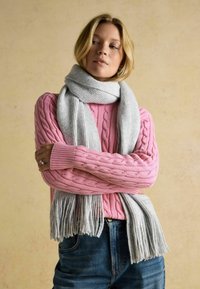Ružový sveter s vrkočovým vzorom a rebrovanými manžetami, doplnený svetlosivým šálom s třásňami. Neformálne rifľové nohavice pridávajú outfitu textúru.