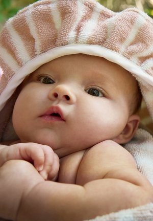 Blød pink og hvid stribet håndklæde på en babys hoved, som afslører runde kinder, store øjne og hænderne samlet mod kroppen.