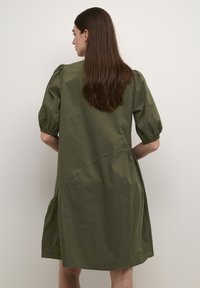 Olive zaļa, līdz ceļiem garuma kleita ar pūkainām piedurknēm, sānu šuvju detaļām un saplacinātu apakšmalu. Izgatavota no viegla auduma, gluda tekstūra.