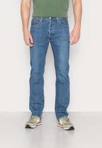 Homme portant un jean bleu en denim, un t-shirt vert et des baskets beiges debout sur un sol gris devant un fond blanc.