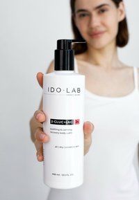 Biała cylindryczna butelka z czarną pompką. Etykieta zawiera napisy "IDO•LAB" i "β-GLUC+cAG 2%". Na etykiecie umieszczono tekst "łagodzący i kojący" oraz "wrażliwa skóra".