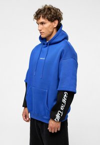 Blauwe hoodie met een voorzak en verstelbare koord, met de tekst "IMPACT" op de borst en "CREATIVE CANG" op de zwarte mouwen.