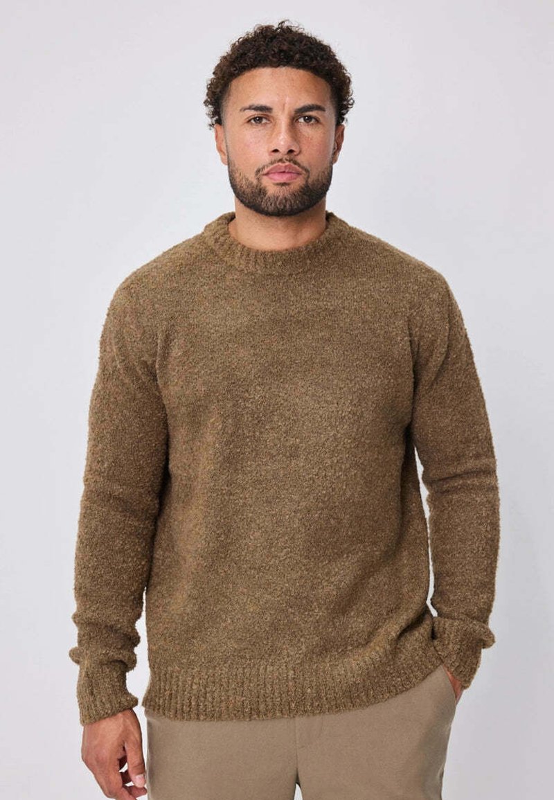 Homme aux cheveux courts et bouclés et à la barbe, portant un pull marron texturé et un pantalon beige, debout avec une main dans la poche.