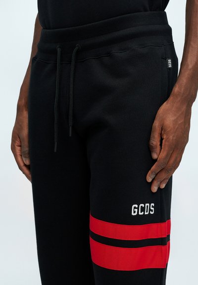 Svarta sweatshirtbyxor med röda ränder på sidan, med en vit broderad "GCDS"-logotyp. Tillverkade av strukturerat material med dragsko i midjan.