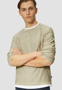 Beige langlangshirt met zachte, gestructureerde stof. Heeft een ronde halslijn en opgerolde mouwen. Gelaagd over een witte basis shirt.