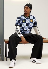 Maillot à manches courtes bleu et noir à motifs avec des accents blancs, porté avec un pantalon large noir et des chaussures blanches ornées de rayures noires.