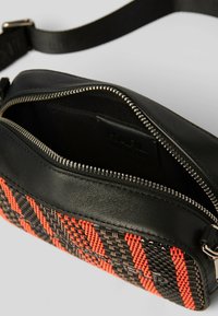 Zwarte leren tas met een geweven rood en zwart patroon, ritssluiting en verstelbare schouderband. Binnenkant is zwart, met een stoffen voering.