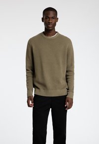 Olive zaļas adītas džemperis ar teksturētu rakstu, ribotām piedurknēm un apkakli, apvienots ar melnām biksēm, modelis stāv baltā fona priekšā.