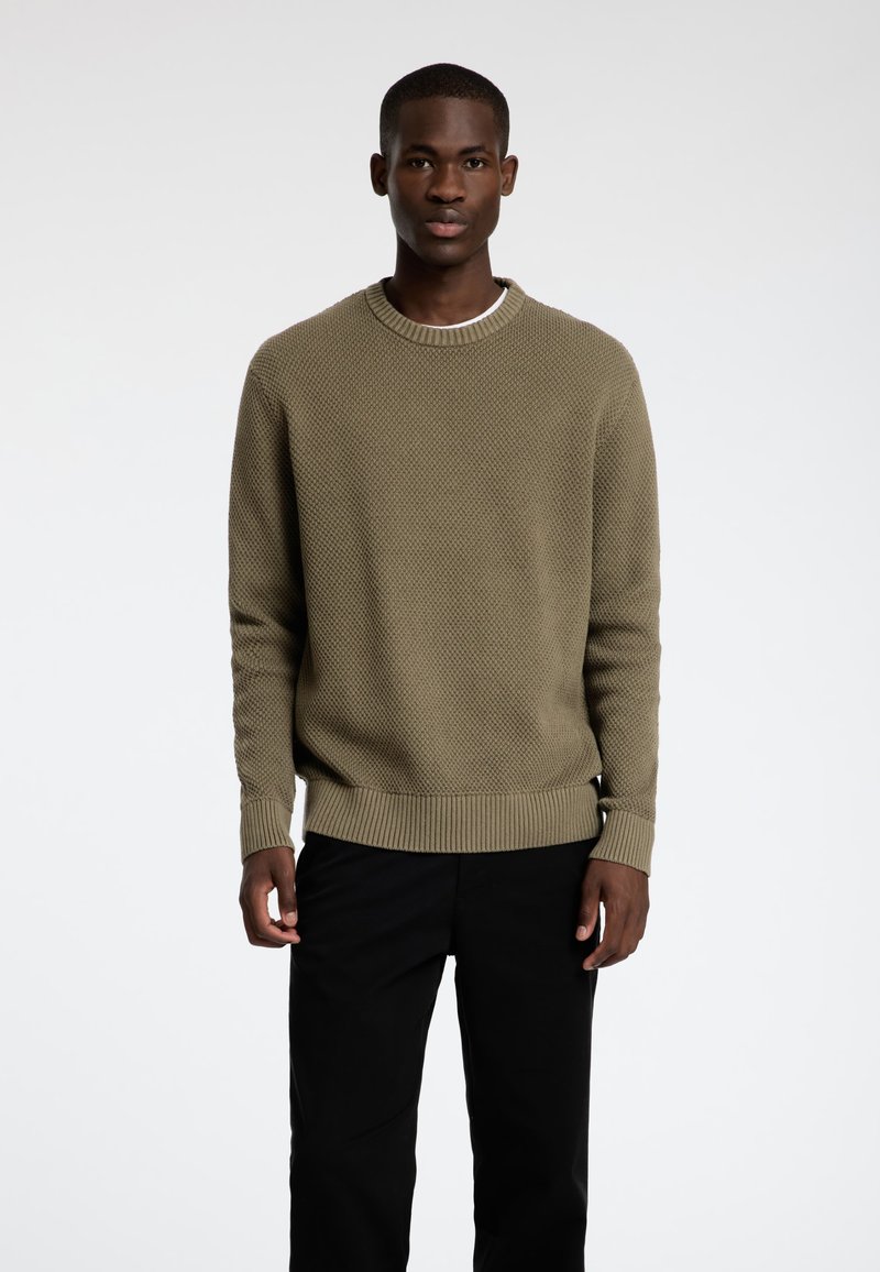 Olive zaļas adītas džemperis ar teksturētu rakstu, ribotām piedurknēm un apkakli, apvienots ar melnām biksēm, modelis stāv baltā fona priekšā.