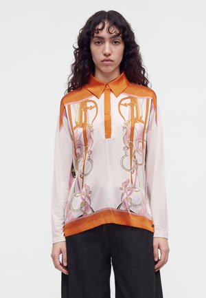 Femme aux longs cheveux bouclés porte un chemisier blanc à manches longues avec un col orange et des motifs floraux, anneaux et harnais, associé à un pantalon noir.