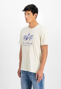 Krémové bavlněné tričko s modro-bílým grafickým logem se pruhy a textem "ALPHA INDUSTRIES." Krátké rukávy, uvolněný střih.