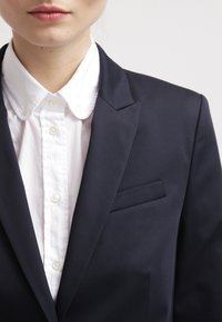 Persona vestida con una camisa blanca de botones y un blazer azul marino oscuro, recortada para mostrar la parte inferior de la cara y la parte superior del torso contra un fondo claro.