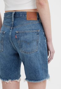 Shorts en denim bleu clair avec un ourlet effiloché, dotés d'une patch en cuir marron à l'arrière et d'une étiquette rouge emblématique sur la poche gauche.