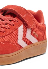 Czerwona zamszowa i siateczkowa sportowa tenisówka z paskiem na rzep, różowymi akcentami i teksturowaną gumową podeszwą w kolorze brązowym. Zawiera elementy w kształcie zygzaka.