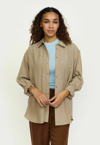 Beige knap-up skjorte med afslappet pasform, lange ærmer og opkrøllede manchetter, båret over en lyseblå tætsiddende top og parret med brune bukser.