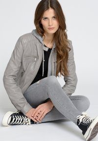 Grijze leren jas met capuchon, zwarte top, grijze skinny jeans en zwarte hoge sneakers. Toont ritsen en een verouderd jeansdetail.