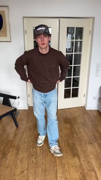 Νέος άνδρας φορώντας μαύρο καπέλο, καφέ φούτερ, ανοιχτό μπλε τζιν και λευκά αθλητικά παπούτσια, στέκεται σε ξύλινο δάπεδο μπροστά από πόρτες με γυάλινα πάνελ.