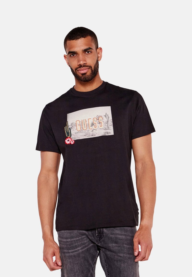 Camiseta de algodón negra con un gráfico rectangular que muestra la palabra "GUESS", cactus y flores. Cuello redondo y mangas cortas. Corte regular.