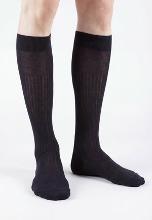 Beine mit kniehohen schwarzen, gerippten Kompressionssocken stehend auf einer weißen Fläche.