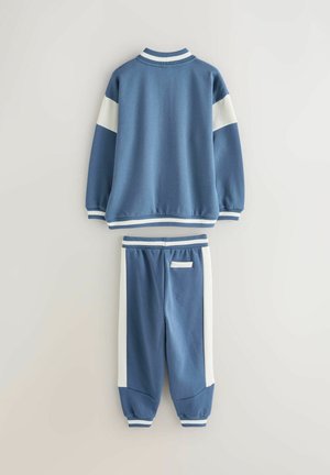 Blåt sæt med sweatshirt og matchende bukser, lavet af bomuldsblanding. Har hvide sidestriber, ribstrikkede manchetter og rund hals.