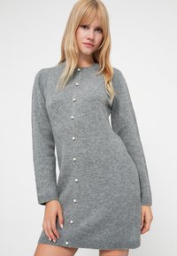 Γκρι πλεκτό φόρεμα με στρογγυλή λαιμόκοψη, μακριά μανίκια και κουμπιά κατά μήκος του μπροστινού μέρους, με πετρώματα μαργαριταριών να τρέχουν κάθετα στο κέντρο.