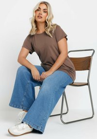 Brun bomulds-t-shirt med korte ærmer parret med brede blå denimbukser. Model siddende på en metalrammet træsæde, iført hvide sneakers.