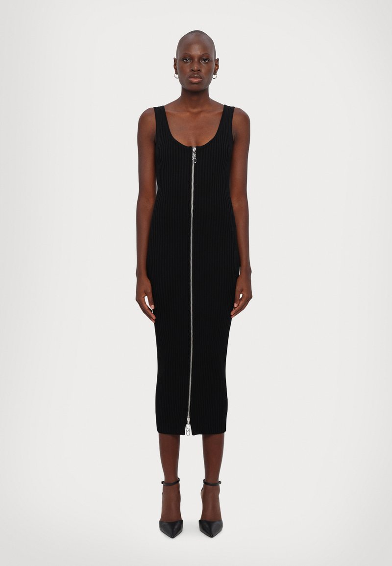 Robe noire en tricot côtelé avec une longueur mi-mollet, sans manches, dotée d'une fermeture éclair sur le devant et d'une silhouette ajustée. Chaussures à talons hauts noires.