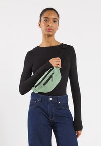 Sac bandoulière en velours côtelé vert avec deux poches zippées noires, porté en travers du corps. Mannequin en chemise noire à manches longues et jean bleu.
